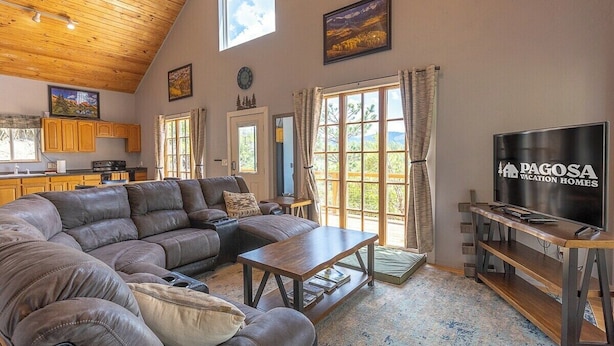 A spacious living room with a large sectional sofa, a wooden coffee table, and a television. There is a kitchen area with wooden cabinets and a window with curtains. - Hot Tub, Secluded, Borders National Forest, New AC (Pagosa Springs)