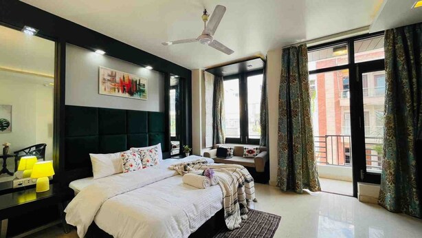 A modern hotel bedroom with a large bed, a sofa, a desk, and a ceiling fan.