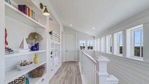 Un long couloir étroit aux murs en bois peints en blanc et au plancher en bois. Des étagères sont disposées le long d’un des murs et contiennent des livres, des objets décoratifs et des paniers. L’autre côté est agrémenté d’une rambarde blanche et de grandes fenêtres.