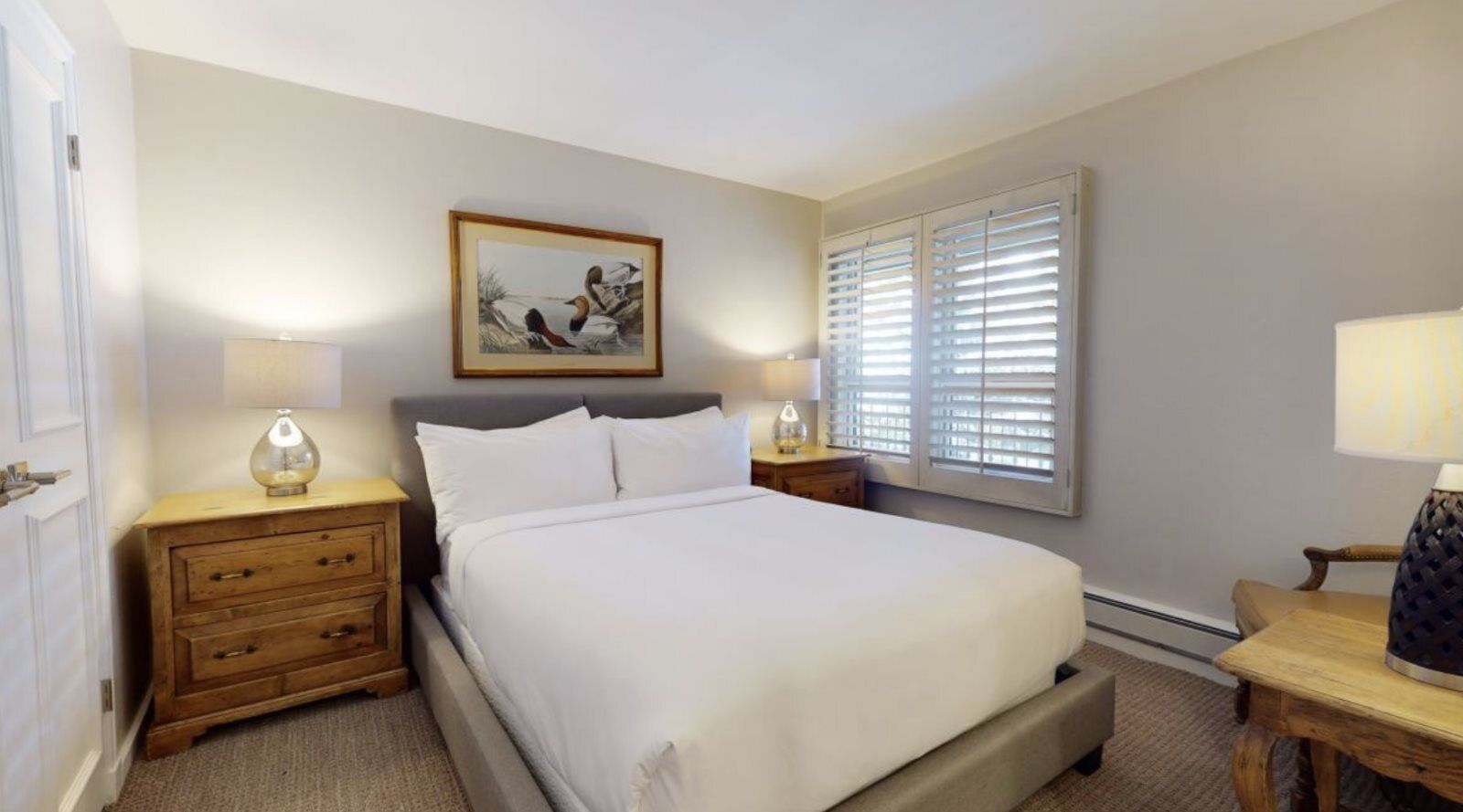 A neatly arranged bedroom with a bed, bedside lamps, a wooden nightstand, a framed picture on the wall, and a window with blinds.