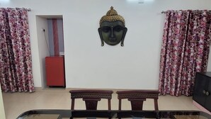 A room with a glass-top table, two wooden chairs, a Buddha head sculpture on the wall, and floral curtains. - Dhairya Villa Home Away From Home (Jammu)