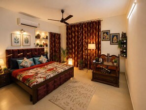 Un dormitorio con cama, mesitas de noche, escritorio, silla, ventilador de techo, cortinas, aire acondicionado instalado en la pared, una obra de arte enmarcada y una planta en maceta.