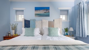 Un dormitorio con cama, un cuadro de un pueblo costero, dos ventanas, una mesita de noche con lámpara y una ventana con cortinas estampadas de estrellas.