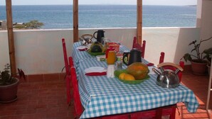 Een gedekte tafel op een balkon, met uitzicht op de zee, een geruit tafelkleed, verschillende gerechten en een waterkoker.
