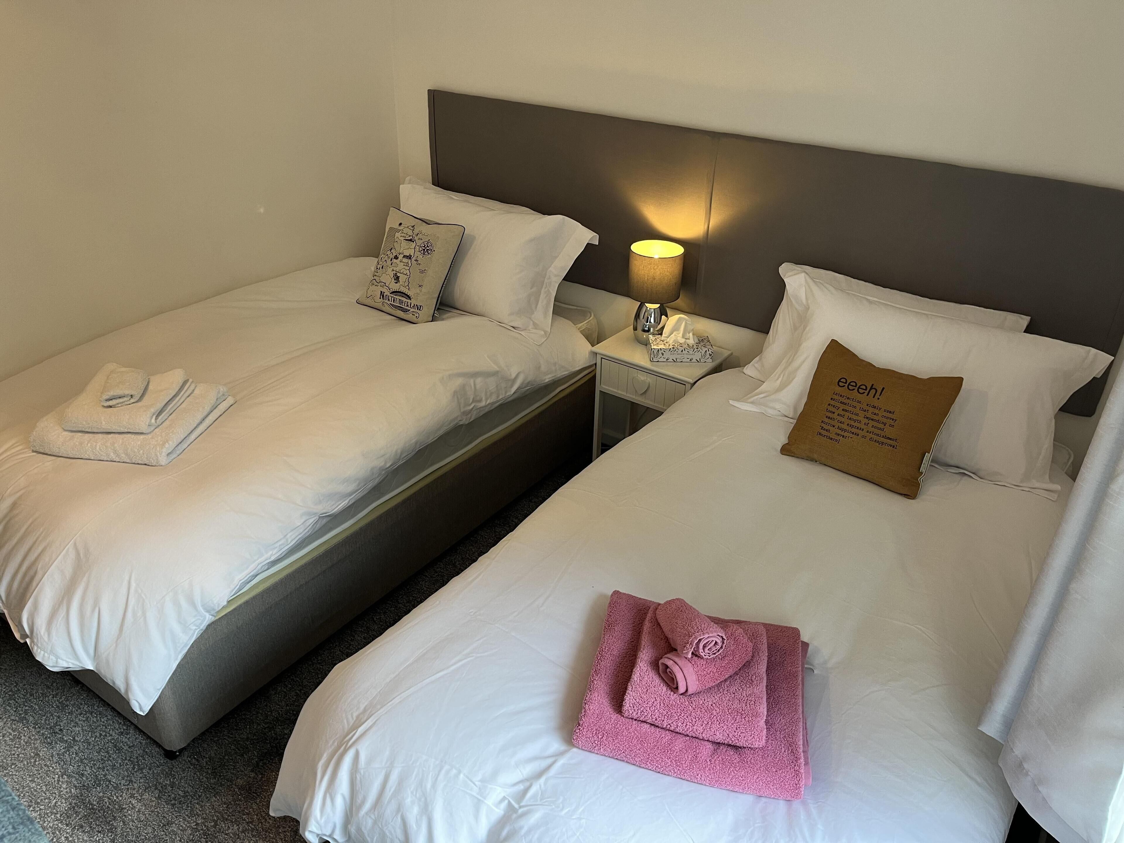 Deux lits simples avec du linge de lit blanc, une table de chevet avec une lampe et un oreiller marron orné d’un motif.