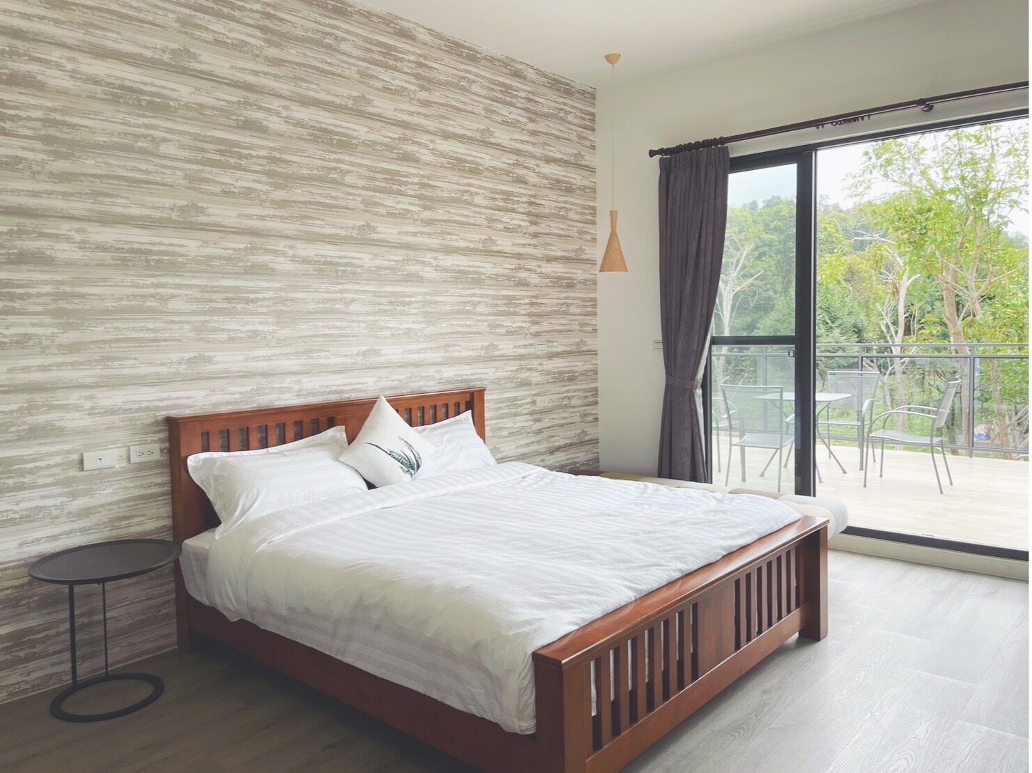 Une chambre moderne avec un lit en bois, une table de chevet ronde et une grande fenêtre donnant sur un paysage verdoyant.