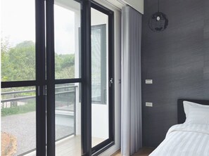 A modern hotel room with a large sliding glass door leading to a balcony, a bed with white linens, and a dark textured wall.
