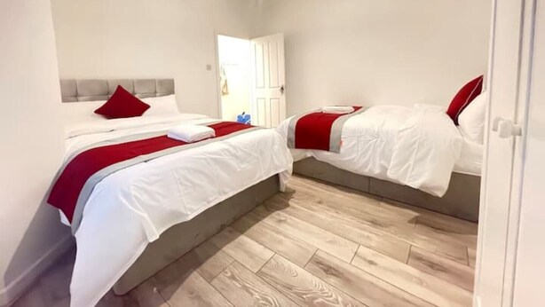A hotel room with two beds, each with white bedding and red accents, a wooden floor, and a white door. - Coast View Professional let (Whitley Bay)