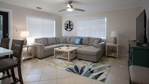 A living room with a sectional sofa, a coffee table, a TV, and a ceiling fan. - Tranquil Retreat by the Beach-4 bedroom 3 bath home w/private pool, parking (Key West)