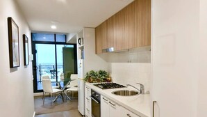 Una cocina moderna con armarios blancos, un lavamanos de acero inoxidable y una estufa a gas. Se ve un comedor con una mesa de vidrio y sillas a través de una puerta corrediza de cristal.