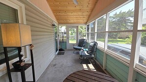 A covered porch with a wooden ceiling, a table with a lamp, and two chairs. - Dunn In the Sun: Pet Friendly with large fenced yard. (Ocracoke)