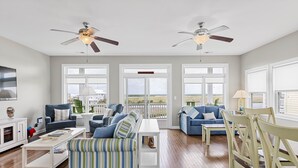 Amplia sala de estar con dos sofás, una mesa de centro y una mesa de comedor. Cuenta con ventiladores de techo, ventanas grandes y vistas al exterior.