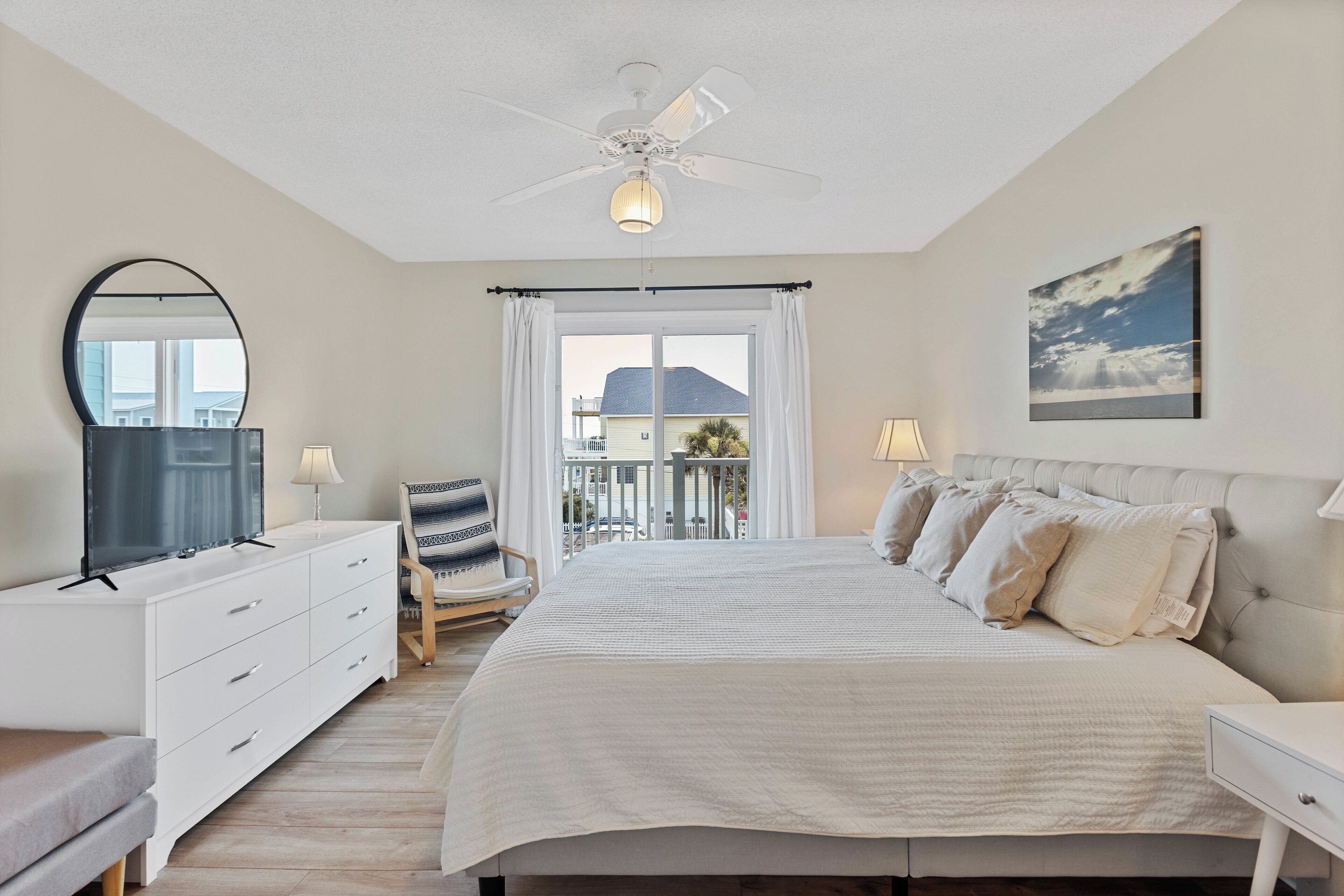 A bedroom with a large bed, a ceiling fan, a window with curtains, a desk with a chair, and a painting on the wall.