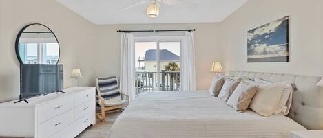 A bedroom with a large bed, a ceiling fan, a window with curtains, a desk with a chair, and a painting on the wall.