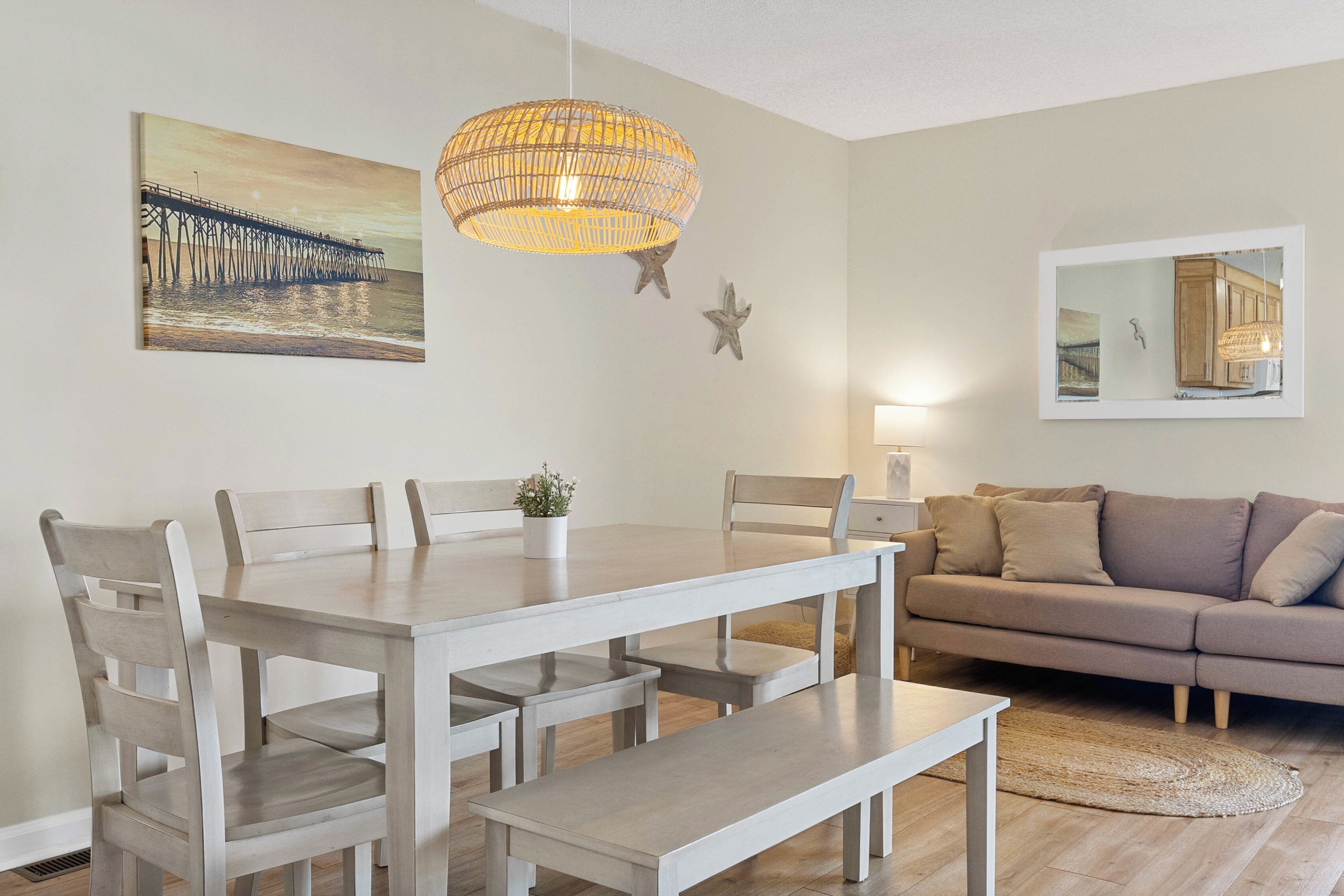 Un comedor con una mesa y sillas de madera, una lámpara colgante y un cuadro de un muelle en la pared.