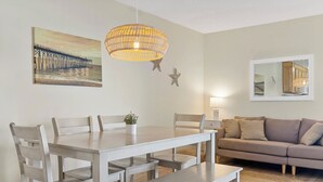 A dining area with a wooden table and chairs, a hanging light fixture, and a painting of a pier on the wall. - Sands V 2B | Ocean Views + Near Area Fun in Carolina Beach (Carolina Beach)