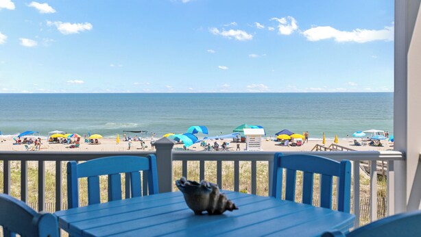 A beach view from a balcony with a table and chairs, umbrellas, and beachgoers in the distance. - Sands V 2B | Ocean Views + Near Area Fun in Carolina Beach (Carolina Beach)