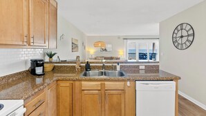 A kitchen with wooden cabinets, a granite countertop, a built-in dishwasher, and a view to a balcony with outdoor seating. - Sands V 2B | Ocean Views + Near Area Fun in Carolina Beach (Carolina Beach)