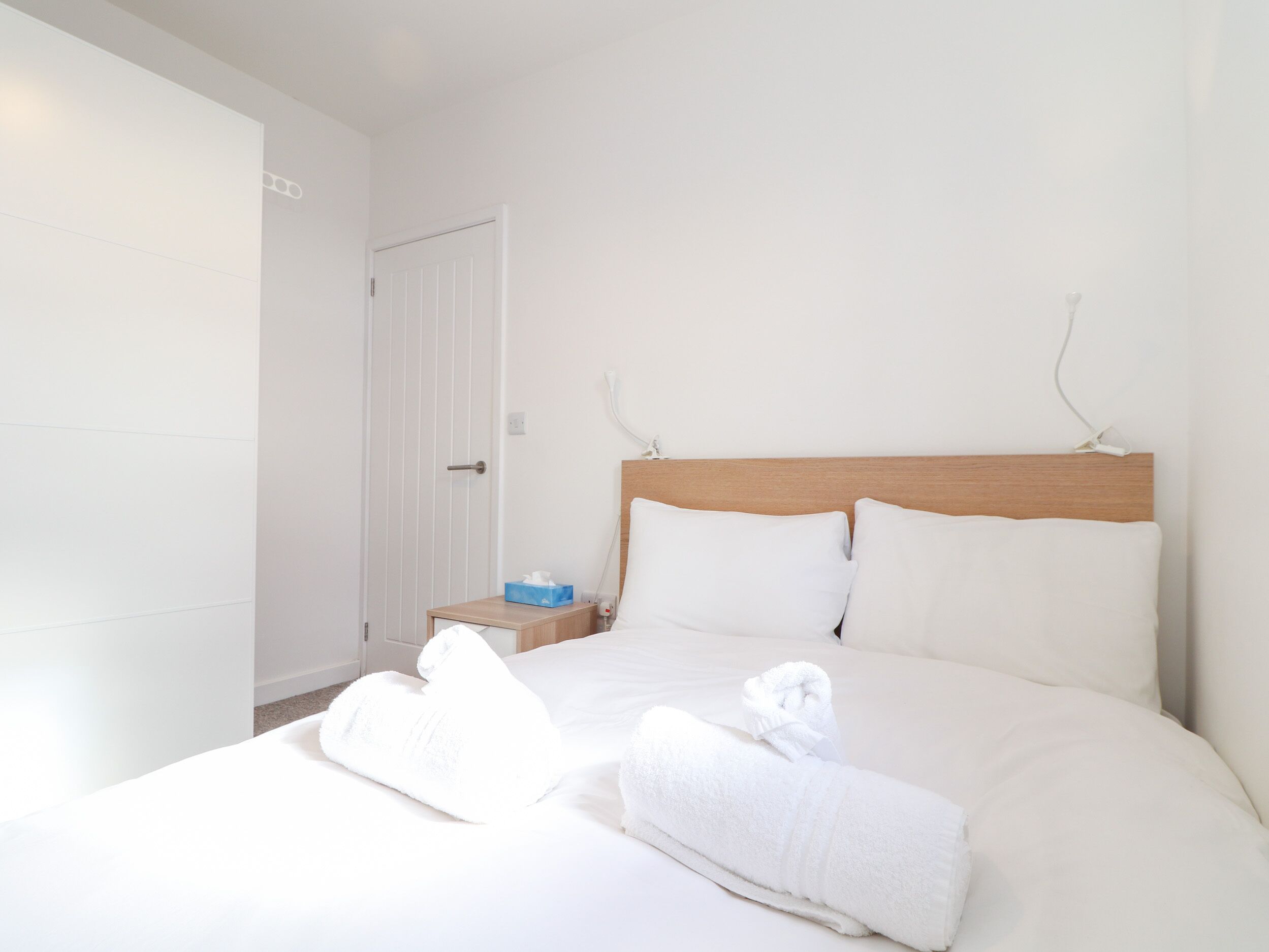 A neatly made bed with white linens and two rolled towels, a wooden headboard, and a bedside table with a tissue box.