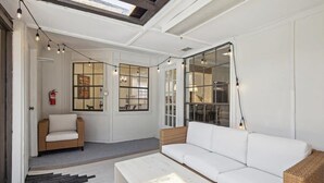 A sunlit room with a white sofa, a wicker chair, and a white coffee table. There are string lights and large windows with glass doors. - Allen Abode - 4 Bedrm. Pool, Fire Pit , Charm (Allen)