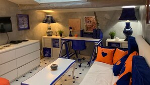A compact room with a skylight, a desk, a chair, a bed with orange pillows, and a small table.