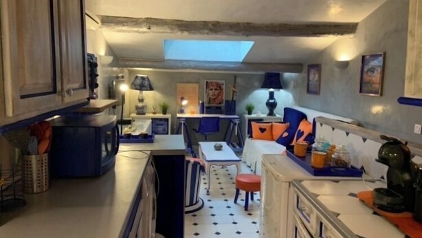 A compact kitchen with a central island, a dining area with a red stool, and a small seating area with a sofa and cushions.