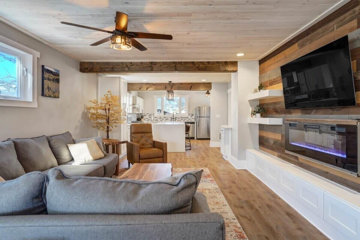 A modern living room with a grey sectional sofa, a ceiling fan, a flat-screen TV mounted on a wooden wall, and a kitchen in the background.