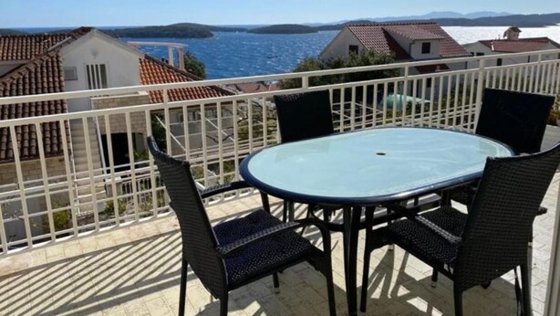 Un balcon doté d’une table et de chaises en verre, offrant une vue sur une zone côtière avec des maisons et une étendue d’eau.