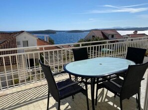 A balcony with a glass table and chairs, overlooking a coastal area with houses and a body of water. - Sunny Mediterranean Apartment Steps from the Beach (Hvar)