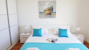 Une chambre d’hôtel avec un lit, deux tables de chevet, une petite table, un tableau représentant un bateau et un magazine posé sur le lit.