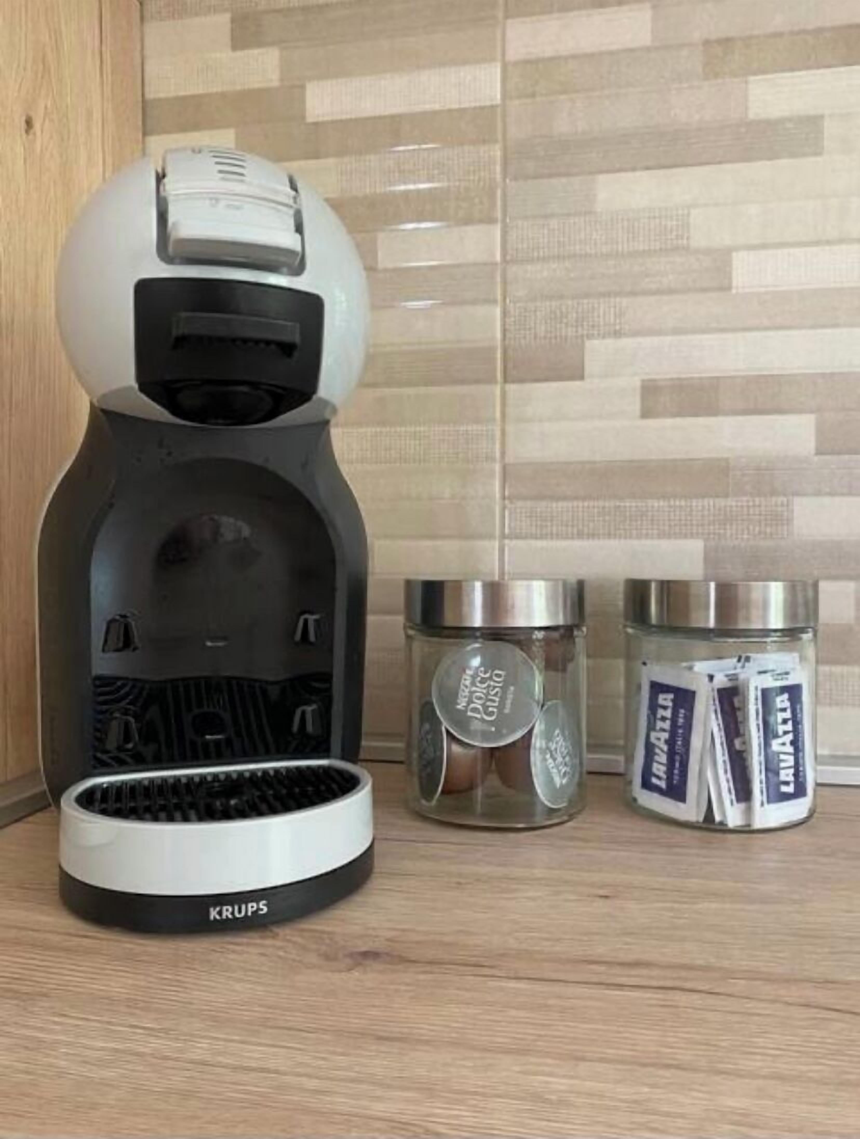 A coffee machine with a Krups brand, two glass containers with coffee pods, and a tiled backsplash.