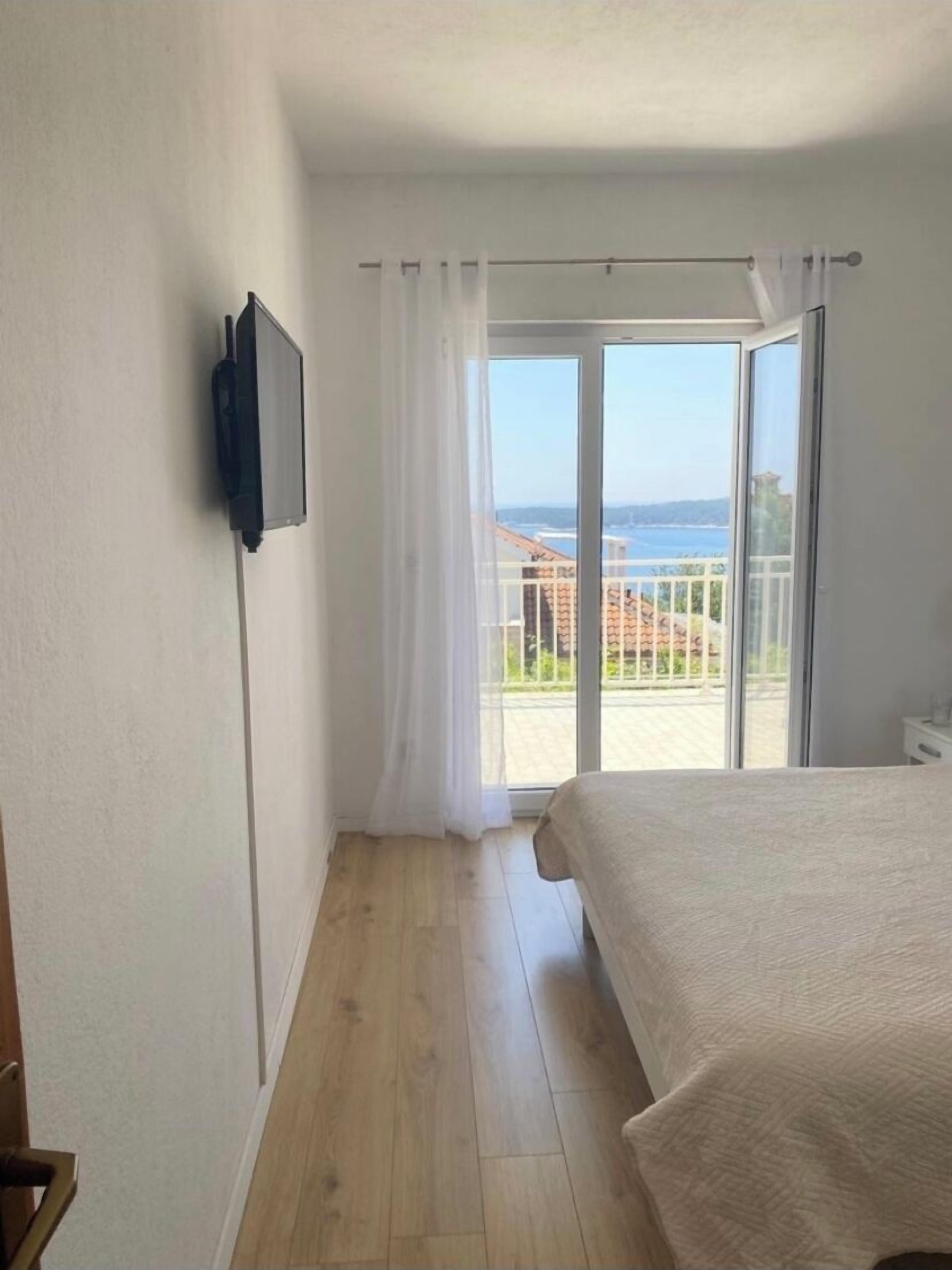 A bedroom with a bed, a TV mounted on the wall, and a balcony with a view of the sea.