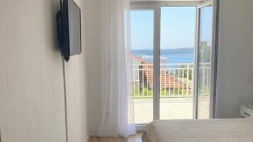 A bedroom with a bed, a TV mounted on the wall, and a balcony with a view of the sea.