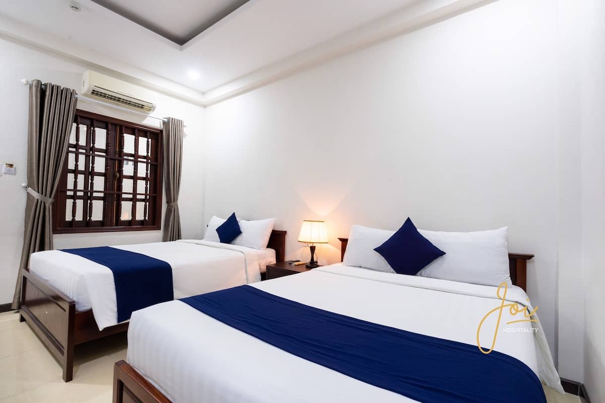 Habitació d'hotel amb dos llits individuals, una tauleta de nit amb una làmpada, un aparell d'aire condicionat i una finestra amb cortines.