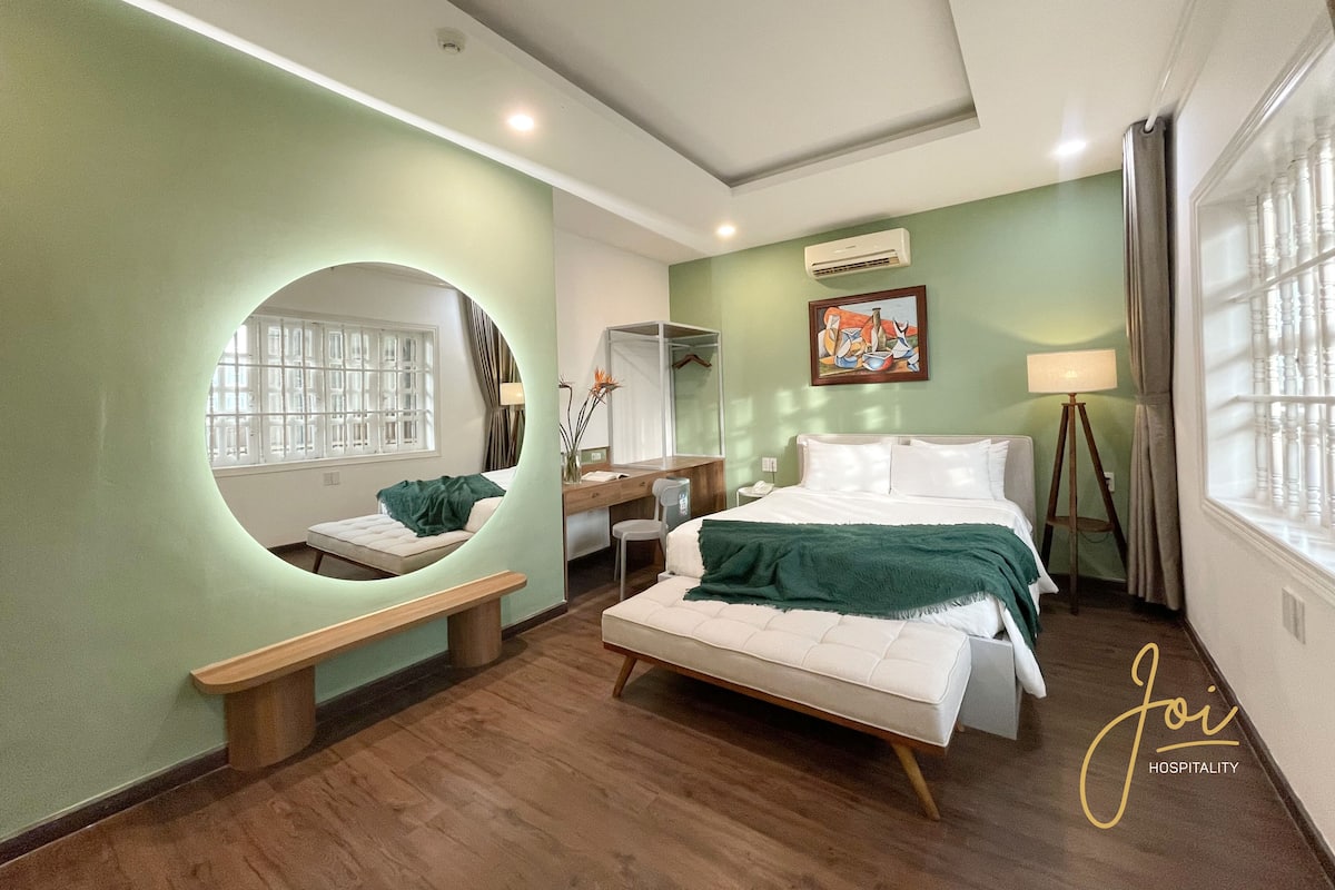Habitació d'hotel amb llit, taula de nit, banqueta, mirall, finestra i obra d'art a la paret.