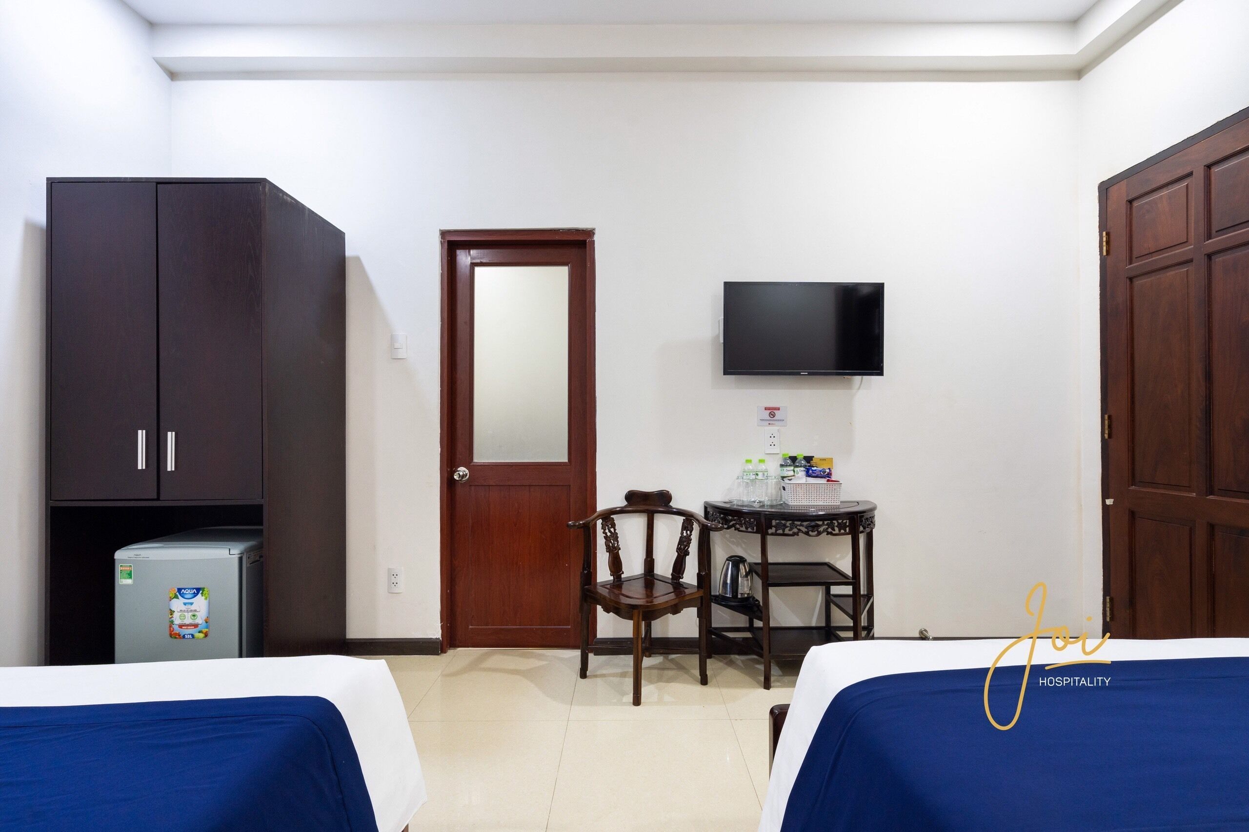 A hotel room with two beds, a small table with a chair, a TV mounted on the wall, a refrigerator, and a wooden wardrobe.