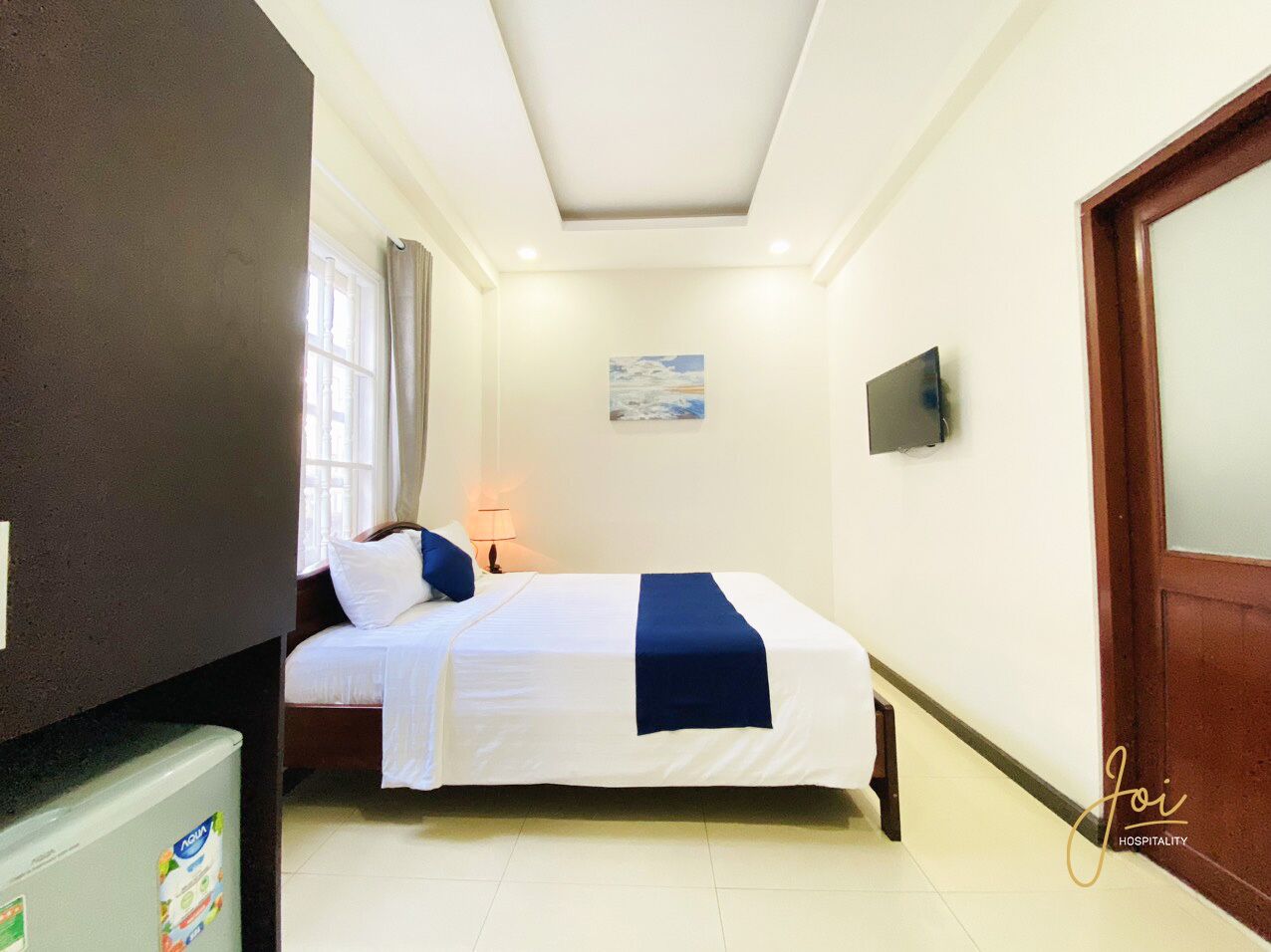 A hotel room with a bed, a TV, a window with curtains, and a door.