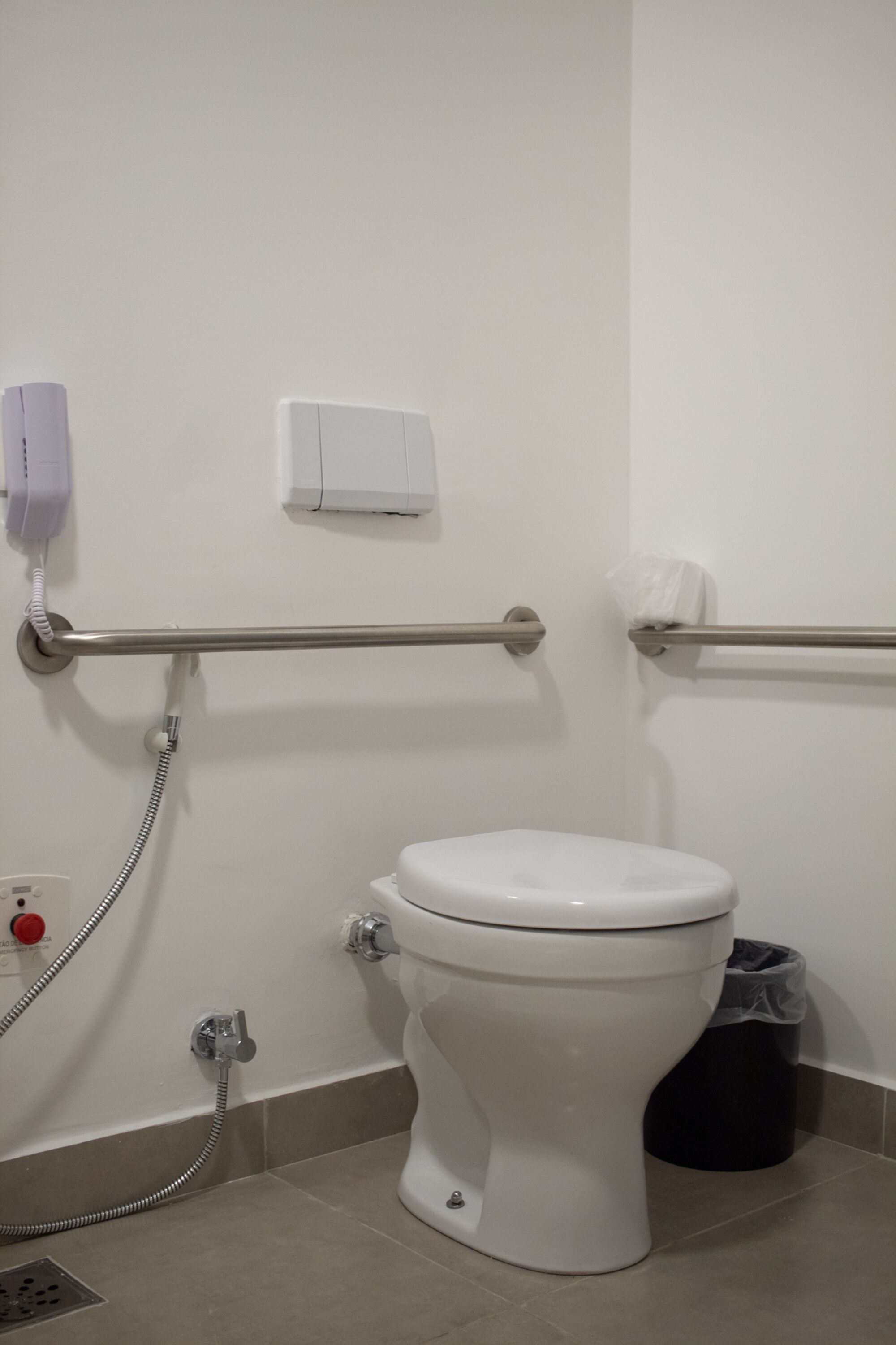 A bathroom with a white toilet, a handrail, a trash bin, and a telephone.