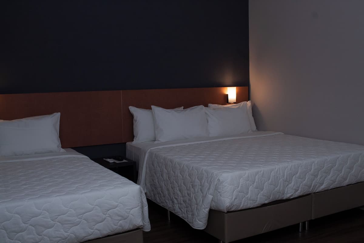 Two single beds with white quilted mattresses and matching pillows, situated against a dark wall with a headboard, and a nightstand with a lamp in between.