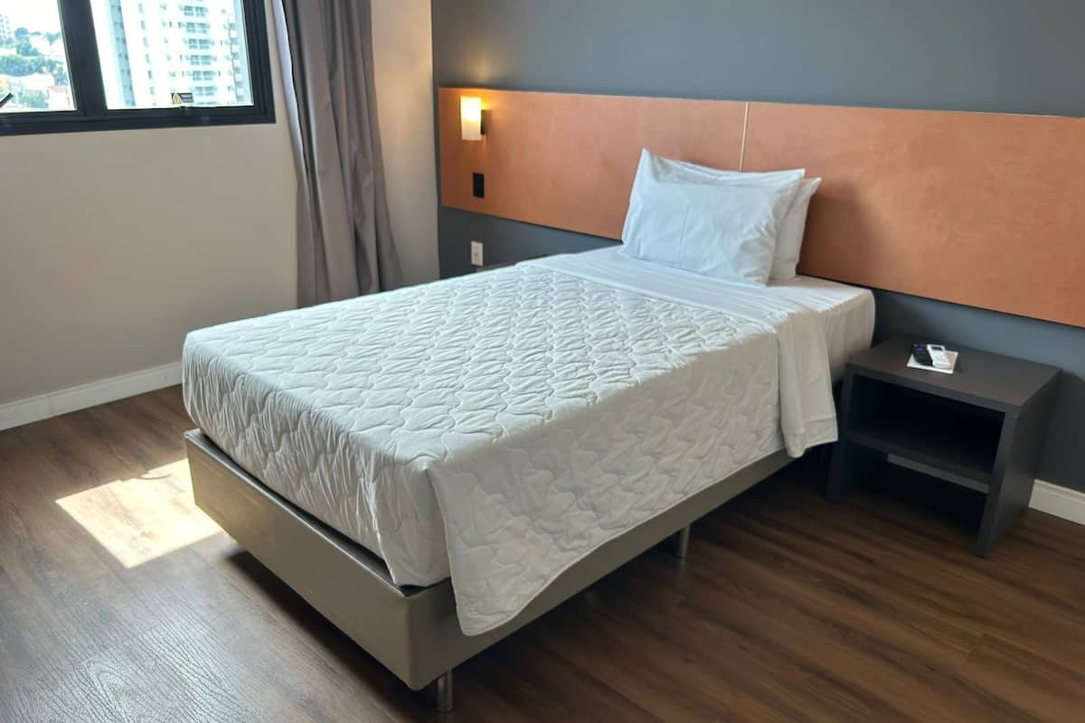 Une chambre d’hôtel moderne, équipée d’un lit, d’une table de chevet, d’une lampe fixée au mur et d’une fenêtre avec des rideaux.