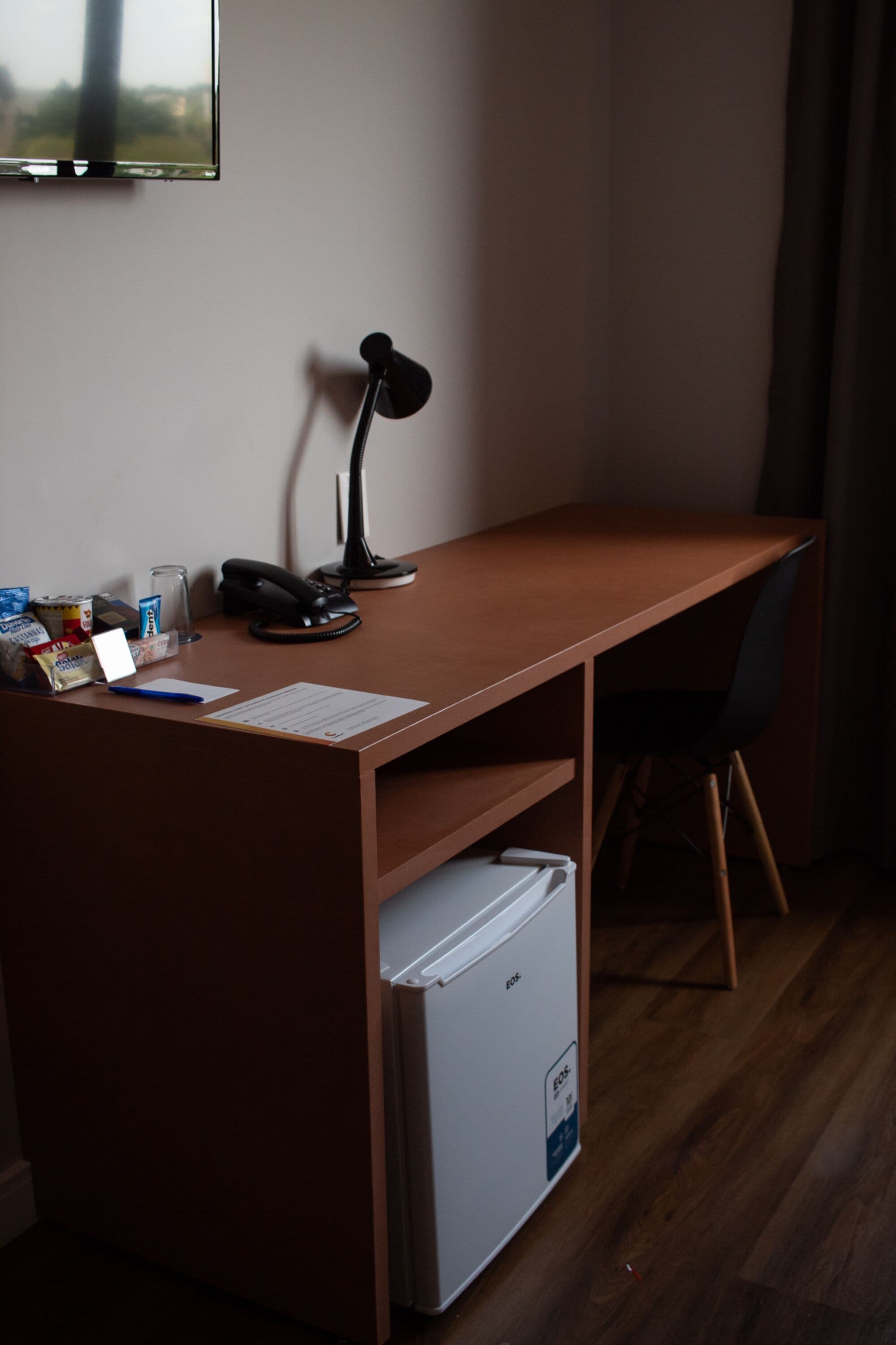 Habitación de hotel con un escritorio de madera, una lámpara de escritorio negra, un mini refrigerador blanco, una silla y una mesita con un vaso y algunos objetos encima.