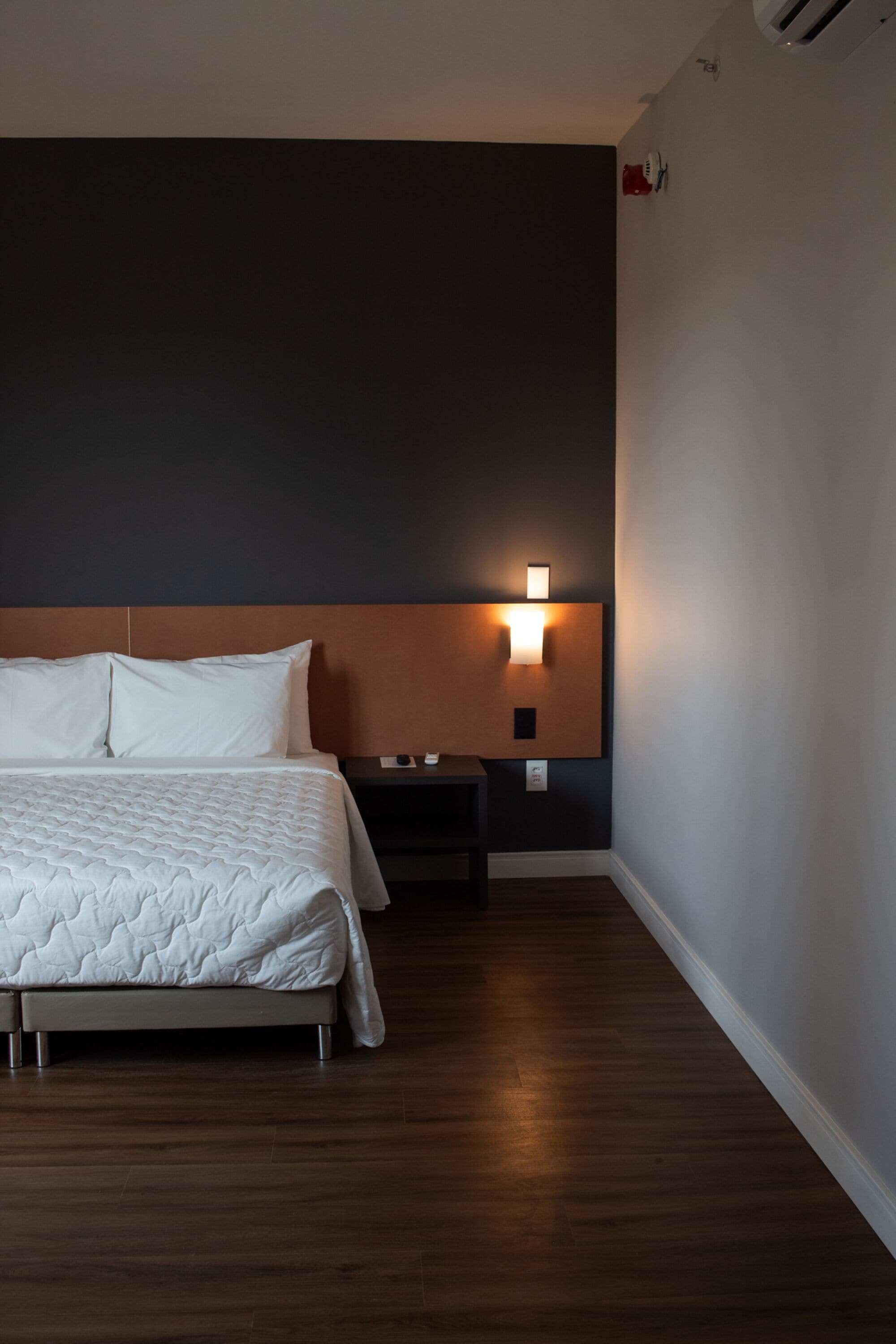 A hotel room with a large bed, a nightstand, a wall-mounted light, and a wooden floor.
