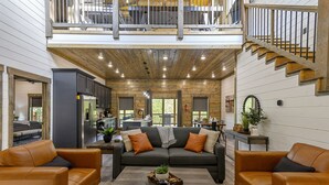 A spacious living area with a high ceiling, a staircase, and a kitchen in the background. - Hilltop Retreat | 3BR Cabin with Mountain Views, Hot Tub & Game Rm | Sleeps 14 (Broken Bow)
