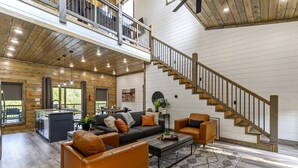 A spacious living area with a wooden staircase, a sofa set, and a kitchen area. - Hilltop Retreat | 3BR Cabin with Mountain Views, Hot Tub & Game Rm | Sleeps 14 (Broken Bow)