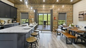 A modern kitchen with granite countertops, wooden cabinets, and a large island with stools. There are two dining areas with wooden tables and benches. - Hilltop Retreat | 3BR Cabin with Mountain Views, Hot Tub & Game Rm | Sleeps 14 (Broken Bow)