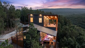 A two-story cabin with a balcony, surrounded by forest. - Hilltop Retreat | 3BR Cabin with Mountain Views, Hot Tub & Game Rm | Sleeps 14 (Broken Bow)
