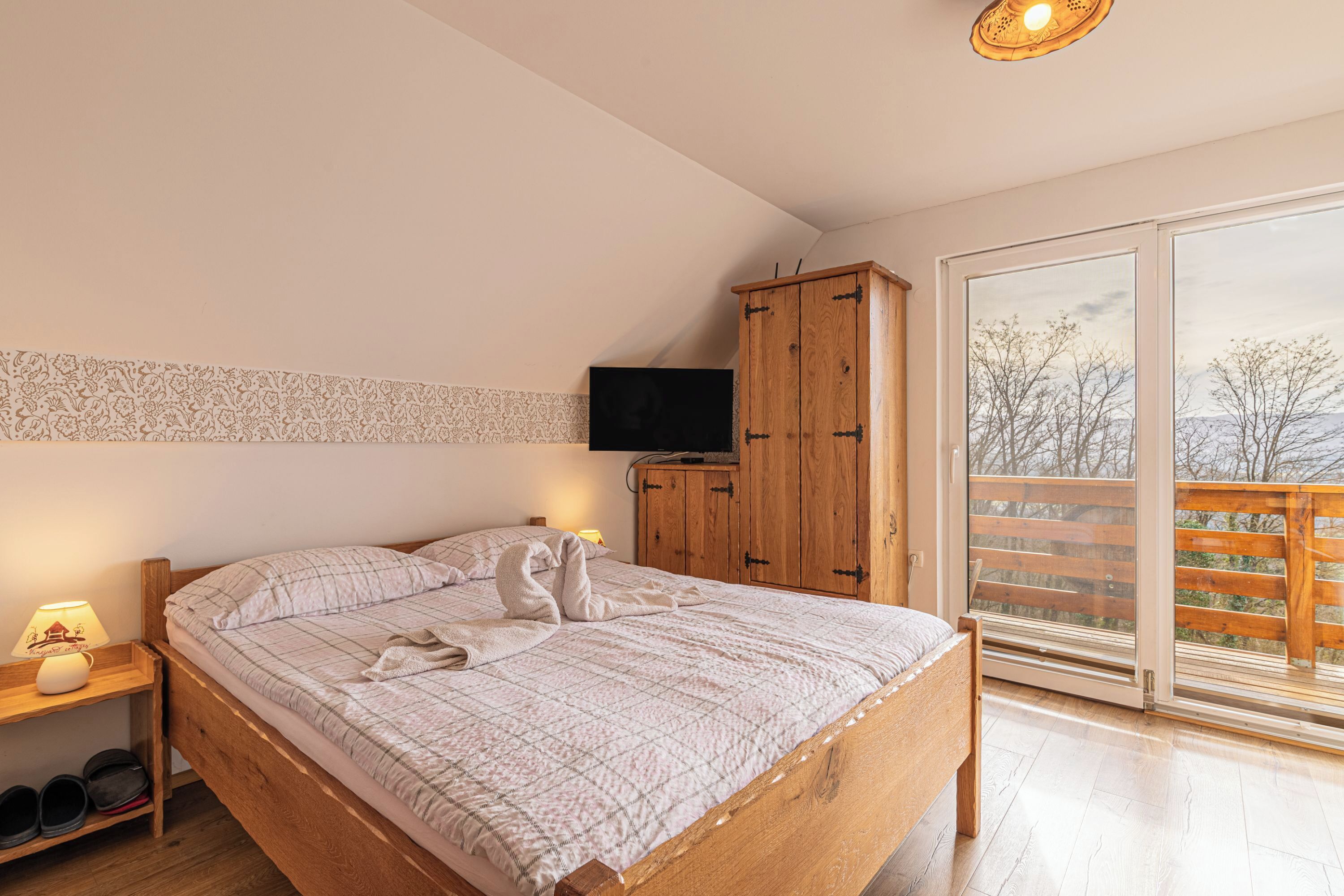 A bedroom with a wooden bed, a television on a wooden cabinet, and a balcony with a view of trees.