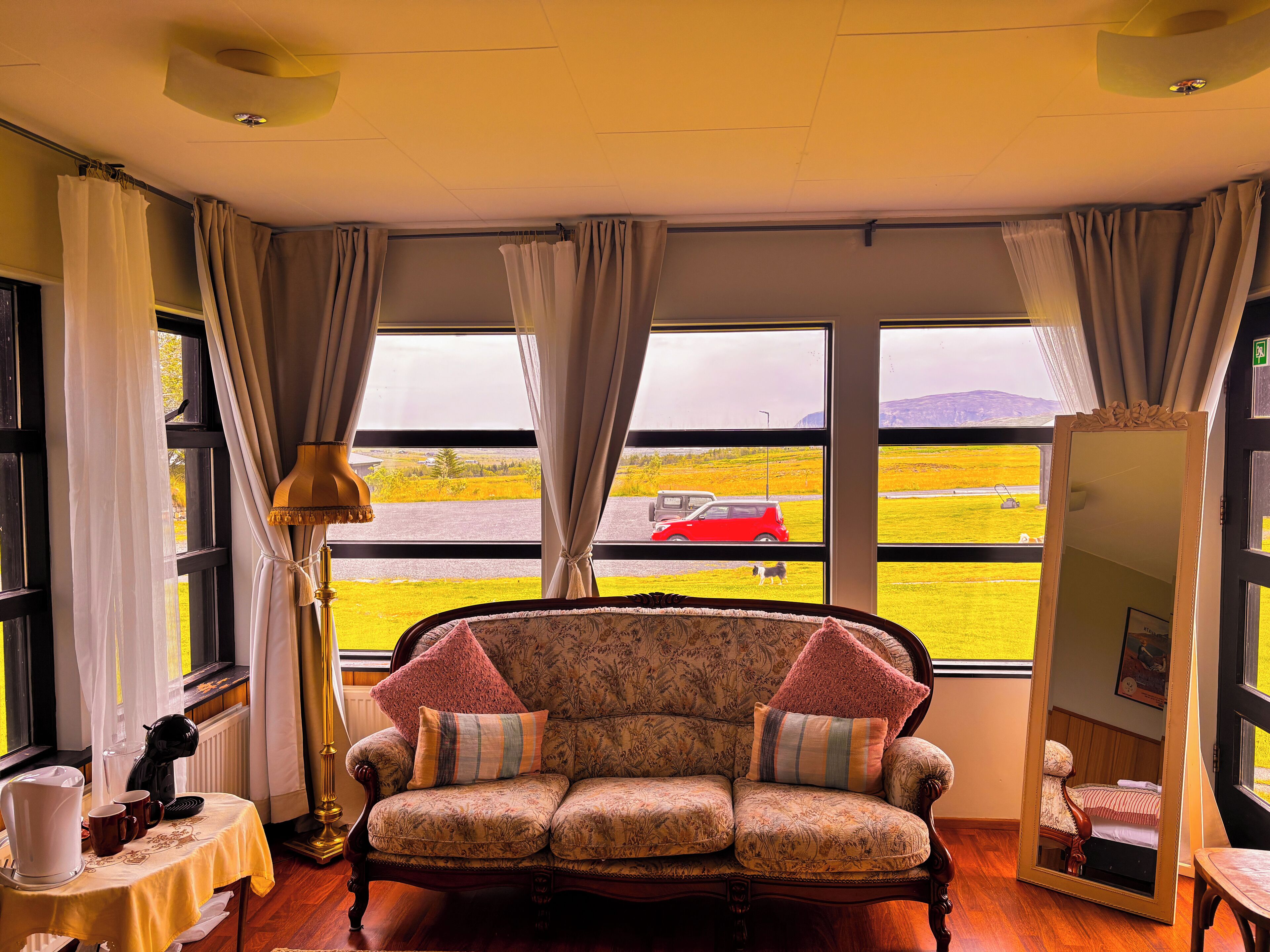 A cozy room with a floral sofa, a table with a lamp, and large windows offering a view of a red car and a dog on a grassy field.