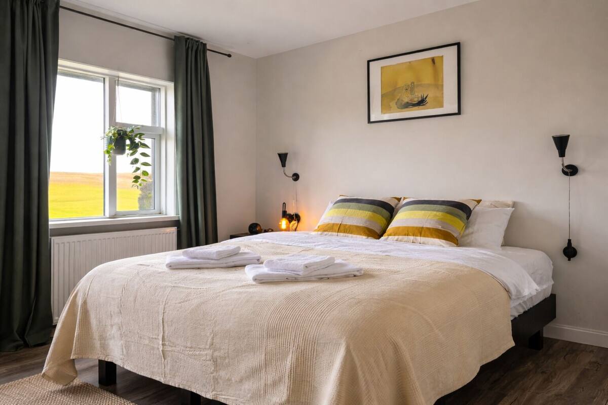 Un lit bien fait, avec du linge de lit blanc et des oreillers à rayures, une œuvre d’art encadrée au-dessus, et une fenêtre donnant sur un champ.
