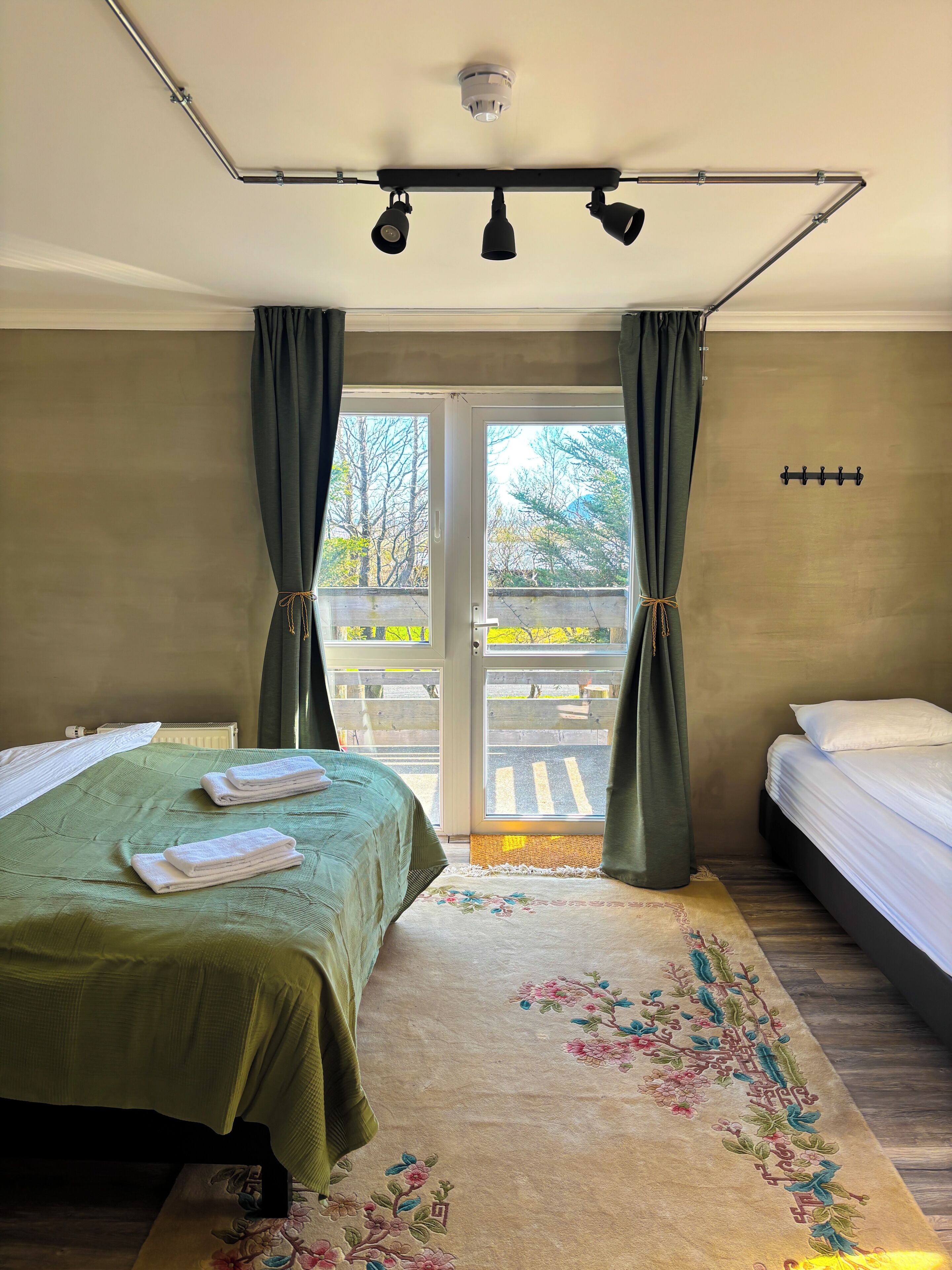 A hotel room with two beds, a green bedspread, a floral rug, a ceiling-mounted light fixture, and a view of a balcony with a railing.